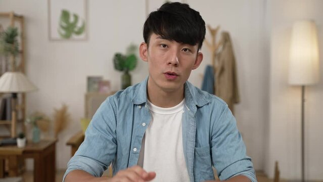 Chinese Asian male creative worker working from home looking and talking to the camera with hand gestures in an online meeting with client through video call