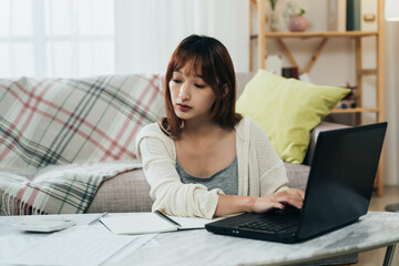 portrait beautiful asian female is looking at the financial document and entering information on the laptop while filing tax online in the living room at home.
