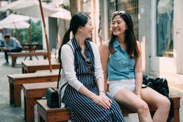 happy asian girls sitting for a rest on a wooden bench are smiling at each other while chatting at...