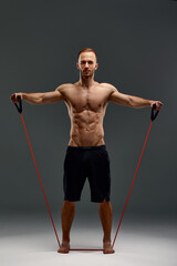 A topless sportsman performs fitness exercises with rubber band in a studio on gray background....