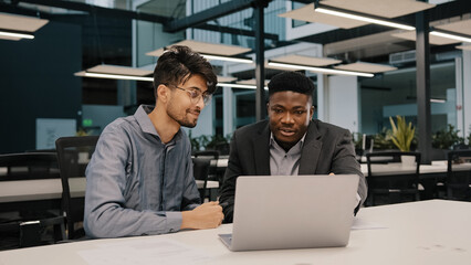 Two multicultural professional male co-workers men colleagues sit at office use laptop discuss...