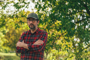 Portrait of male farmer with arms crossed wearing plaid shirt and trucker's hat posing in walnut orchard and looking at camera
