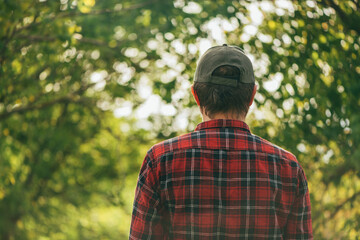 Rear view of male farmer wearing plaid shirt and trucker's hat standing in walnut orchard and looking at trees