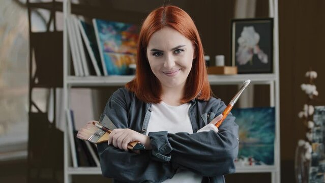 Portrait Caucasian girl confident talented artist woman painter with red hair holding equipment for painting paintbrush trowel spatula crossing arms posing in art studio after drawing pictures design