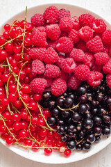 White plate with raspberries, red and black currants on a white background. Sweet healthy vegan food. Top view.