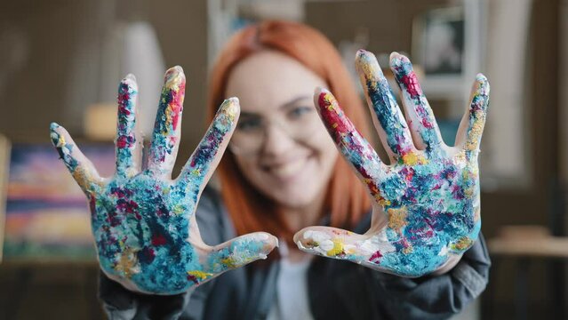 Close-up happy funy woman with red hair painter girl artist in glasses looks at camera stretches forward hands showing dirty palms smeared in acrylic paint laughs inspired by drawing decor design art