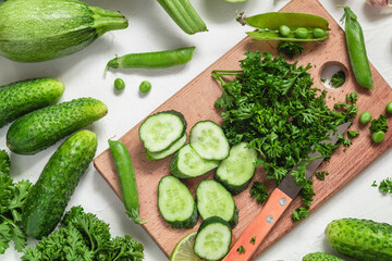 Wooden board with chopped cucumber, parsley, zucchini, garlic. basil. green pea. Assorted green vegetables on a white background. Organic raw produce.