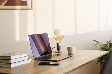Blank screen laptop, cell phone and a stack of books on wooden table. Close up, copy space, white wall background.