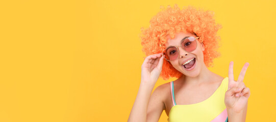 happy kid in sunglasses and swimsuit wearing orange curly wig hair showing peace gesture, peace. Funny teenager child on party, poster banner header with copy space.