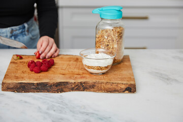 Yogurt with granola, and raspberries and strawberries next to jar with granola in a bright modern kitchen and a woman cutting strawberries