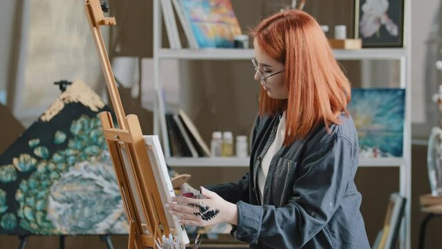 Side view art worker red-haired busy woman in glasses talented artist girl student of painting class in glasses motivated painter create abstract picture with paint brush oil acrylic drawing on canvas