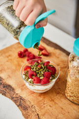A woman topping yogurt, granola, fruit bowl with pumpkin seeds in a bright modern kitchen