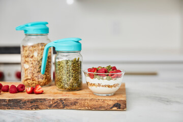 Yogurt with granola topped with raspberries and strawberries next to jars with granola and nuts in a bright modern kitchen
