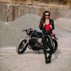 Fototapeta na wymiar beautiful girl with long dark hair red lips in black jacket and black pants standing near black vintage motorcycle in industrial zone