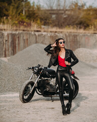 Fototapeta na wymiar beautiful girl with long dark hair red lips in black jacket and black pants standing near black vintage motorcycle in industrial zone
