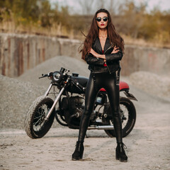 beautiful girl with long dark hair red lips in black jacket and black pants standing near black vintage motorcycle in industrial zone