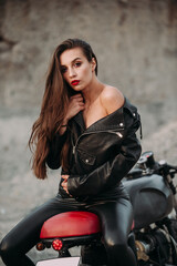 Fototapeta na wymiar beautiful girl with long dark hair red lips in a black jacket and black pants sits on a black vintage motorcycle in an industrial zone