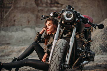 Fototapeta na wymiar beautiful girl with long dark hair red lips in a black jacket and black pants sits near a black vintage motorcycle in an industrial zone