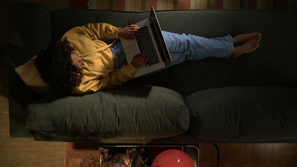 One young black girl lying down on couch opening laptop. Top view angle of person using computer at night © Marco
