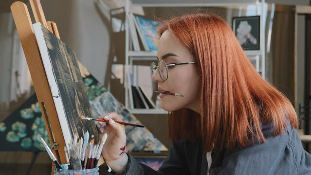 Inspired funny focused artist woman with red hair girl painter wears glasses holds painting brushes in mouth paints with oil paints on canvas on easel in studio inspiration creative beautiful drawing