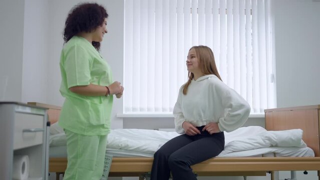 Young woman complaining pain sitting on bed in ward talking to nurse standing in hospital. Caucasian patient and doctor indoors in medical clinic chatting. Illness and treatment concept