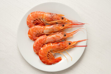 Four cooked red tiger shrimps on a white ceramic plate on white wood from above.