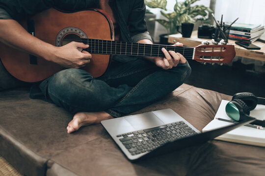 young man relax and playing guitar while sitting on sofa bed in living room at home. Music create melody song, lyrics on laptop and practice concept.