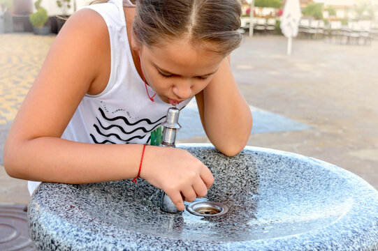 Cute little girl drinks water from public potable fountain faucet fountain on a hot summer day.