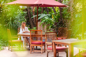 Young smiling woman on tropical vacation in modern luxury resort. Female traveler in bathrobe relax near swimming pool, sitting under a sun umbrella. Poolside spa lounge zone in hotel.