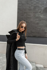 Naklejka premium Fashionable beautiful young woman model with sunglasses in fashionable black clothes with black T-shirt and vintage high waist jeans with coat walking outside