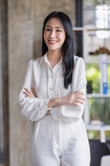 Fototapeta na wymiar Portrait of a smiling young Asian businesswoman leaning against a table while standing working alone in a large modern office