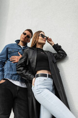 Stylish young beautiful couple in fashionable casual urban clothing with a leather black coat and jeans stand near the wall. Fashionable pretty hipster woman wearing cool vintage fashion sunglasses