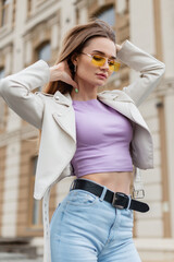 Stylish beautiful young hipster girl with yellow fashion glasses in a trendy white leather jacket...