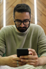 Obraz na płótnie Canvas Young businessman in grey pullover and eyeglasses texting in mobile phone while sitting in cafe or office between two lamps