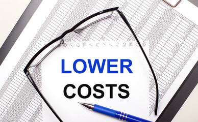 On a light background, a report, black-framed glasses, a pen and a sheet of paper with the text LOWER COSTS. Business concept