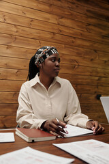 Young serious black woman in white shirt planning work at meeting with colleagues while sitting by table with financial documents