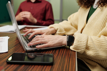 Hands of young businesswoman touching keys of laptop keyboard while sitting by table against male colleague working with papers