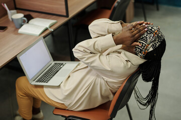 Young very tired businesswoman with laptop on her knees sitting on chair by workplace with her hands on face while having rest