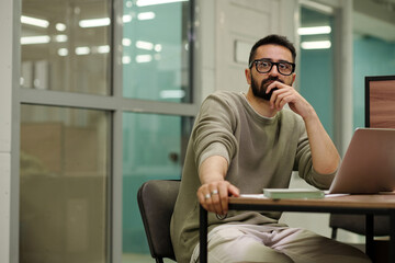 Fototapeta premium Young pensive businessman in eyeglasses sitting by desk in front of laptop and brainstorming while working over new project