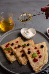 delicious thin pancakes on a gray plate with sour cream and frozen berries in a heart-shaped bowl on a gray background with honey, a woman's hand holds a glass spoon with honey over pancakes