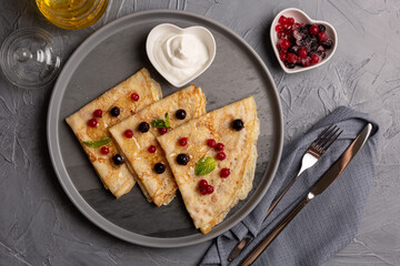 delicious thin pancakes on a gray plate with sour cream and frozen berries in a heart-shaped bowl on a gray background with honey