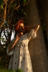 Gorgeous red haired woman in a white dress is posing near the column under sun rays in the park in summer. Warm picture of a beautiful girl under sunlight for poster, wallpaper or background.