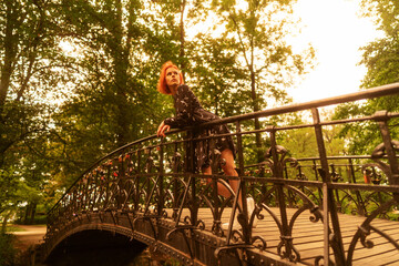 Gorgeous girl walks alone on the bridge over the river in the park under sunny sky. Snapshot of a young pretty woman with red hair in a black dress for cover, poster, wallpaper or background.
