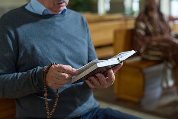 Hands of aged man holding open Bible and wooden rosary beads while reading verses from Gospel or...