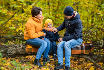 Fototapeta premium Mother, father and little son sit on a plaid on a log in the autumn park and spend time together