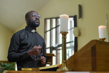 African American priest in black casualwear holding rosary beads during pray while standing by pulpit with two burning candles - obrazy, fototapety, plakaty