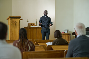 Confident priest of evangelical church with Holy Bible in hand saying sermon while standing in front of intercultural parishioners - obrazy, fototapety, plakaty