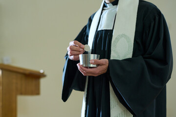 Priest of Catholic church in cassock holding small cup with unleavened bread for communion rite of parishioners during liturgy