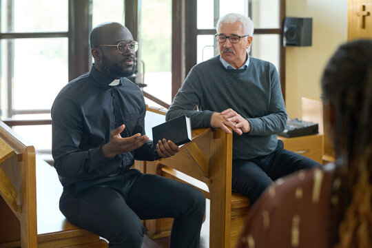 Confident pastor in black shirt with clerical collar explaining verses from Bible to intercultural parishioners of his church after sermon