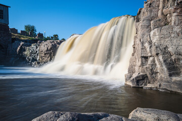 Waterfall with quartzite red rocks at Big Sioux River Park in Sioux Falls, South Dakota. Long exposure photo of the silky flowing water with the view of the Queen Bee Turbine House on the hilltop.